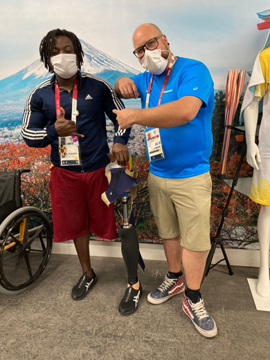 two men wearing face masks standing side by side, one is holding the other's prosthetic leg